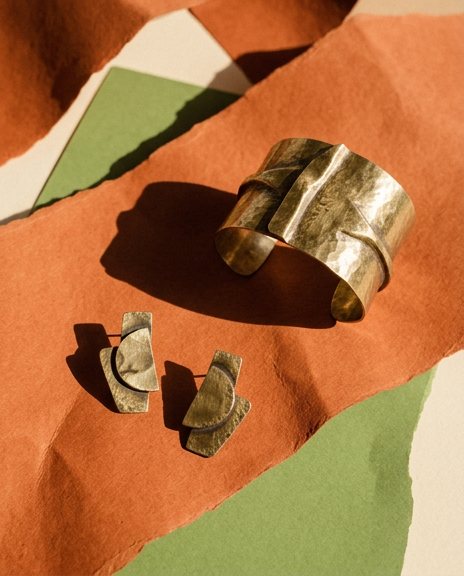 Brass cuff and earrings on torn orange and green paper in hard sunlight